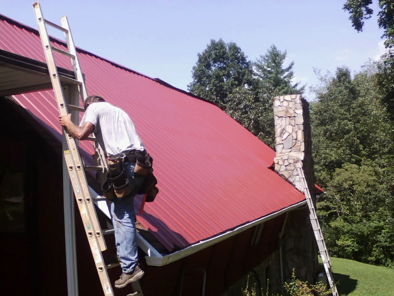 Roofer on a ladder beside a red metal roof and stone chimney — Soffit & Fascia Repair by Roofing Brothers in Sunburg